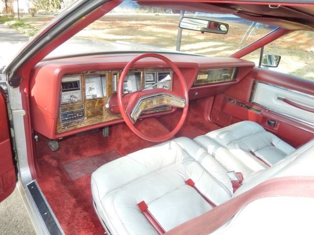 Interior of a vintage car featuring a red dashboard and steering wheel, woodgrain accents, white leather seats, and a red carpet.