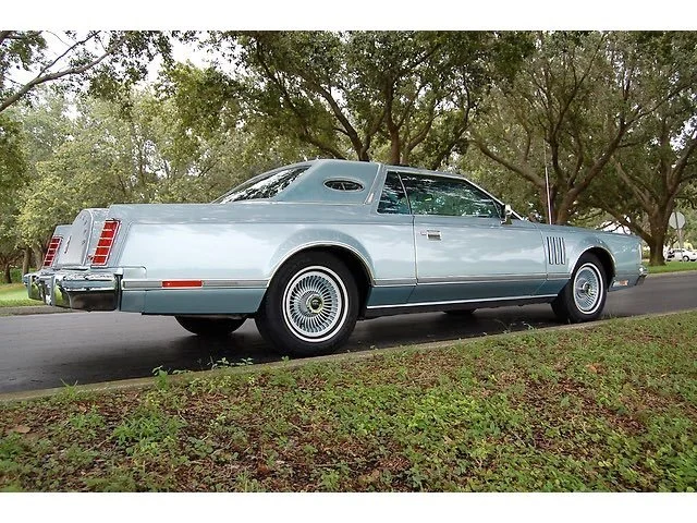 A vintage silver luxury car parked on a street with trees in the background.