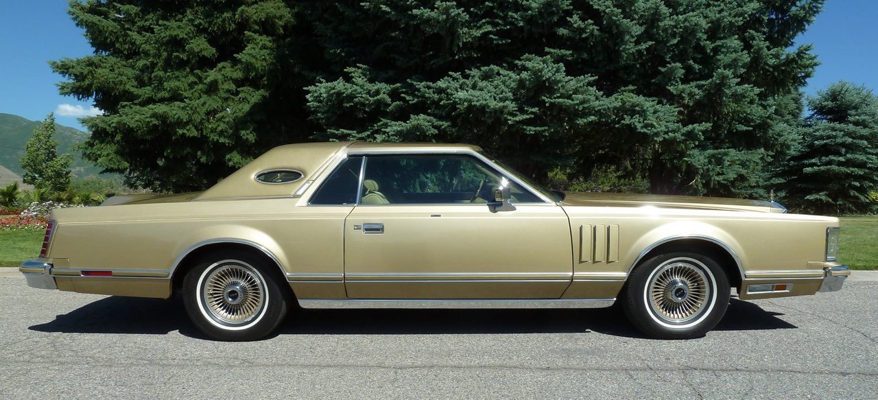 A vintage beige luxury car parked on a street with green trees and hills in the background.