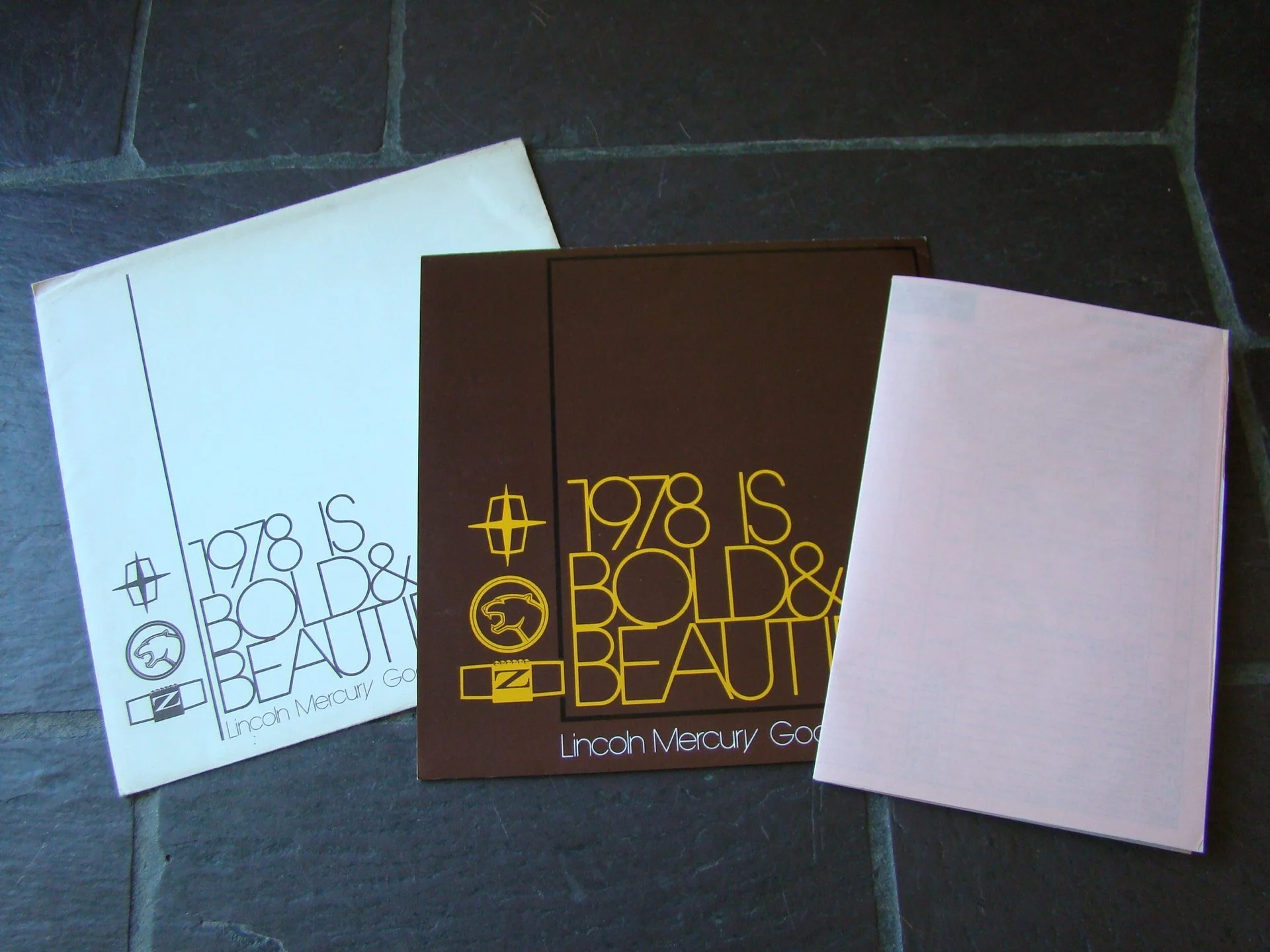 Three booklets or catalogs titled '1978 IS BOLD & BEAUTIFUL' laid out on a dark tiled surface, with one booklet in black and yellow, another in white with black text, and the third mostly blank or with faint lines.