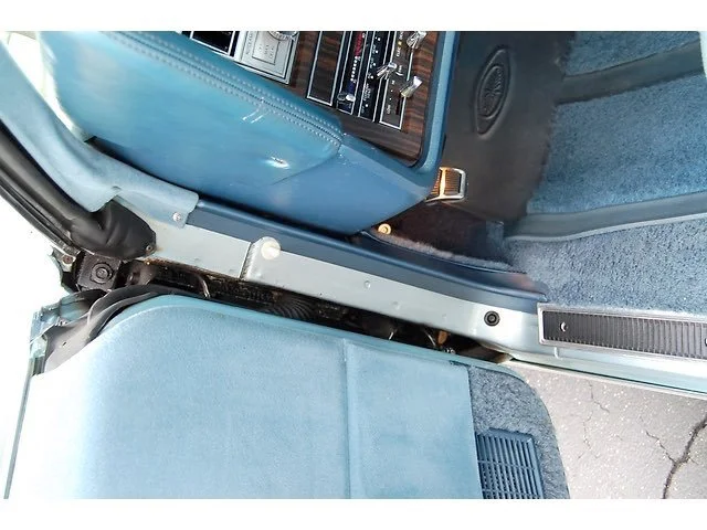 Close-up of the front passenger side interior of a vintage car, showing the dashboard, steering wheel, and part of the seat with a mix of wood, metal, and blue upholstered materials.