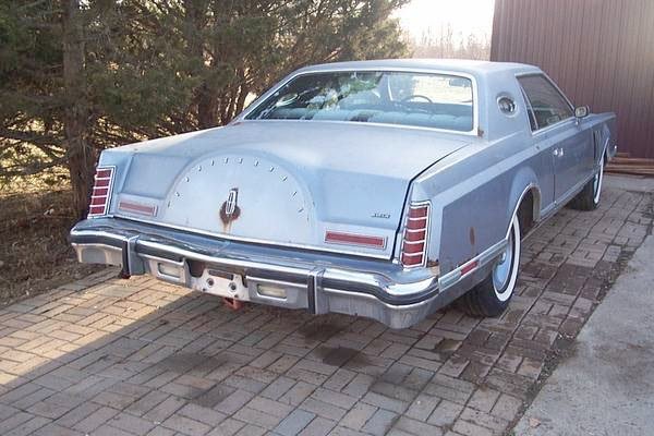 A vintage light blue Cadillac parked on a brick driveway with trees in the background.