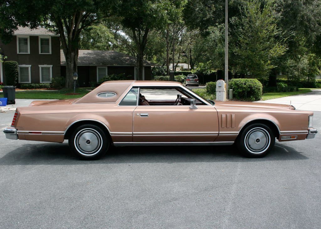 A vintage beige luxury car parked on a suburban street with trees and houses in the background.
