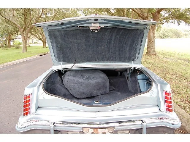 The open trunk of a vintage silver car filled with a black bag and a compact spare tire, parked on a residential street with trees and grass in the background.