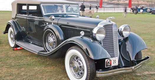 Vintage black luxury car with white-wall tires parked on grass at a car show.
