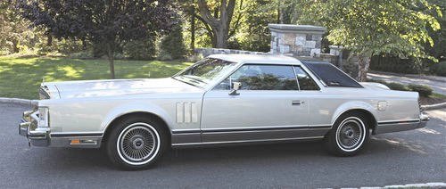 A classic silver 1980s luxury car parked on a street with trees and a stone structure in the background.