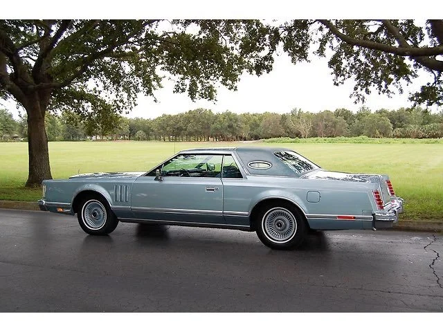 A vintage blue car parked on the side of a road with green grass and trees in the background.