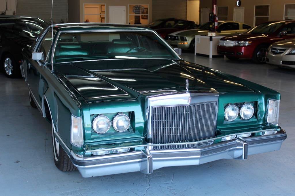 Green vintage luxury car with chrome details parked inside an underground garage, surrounded by other modern cars.