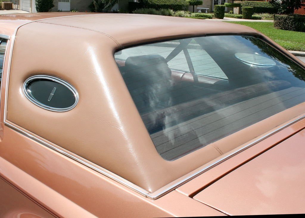 Close-up of the rear window and beige vinyl top of a vintage car with oval-shaped side vent windows.