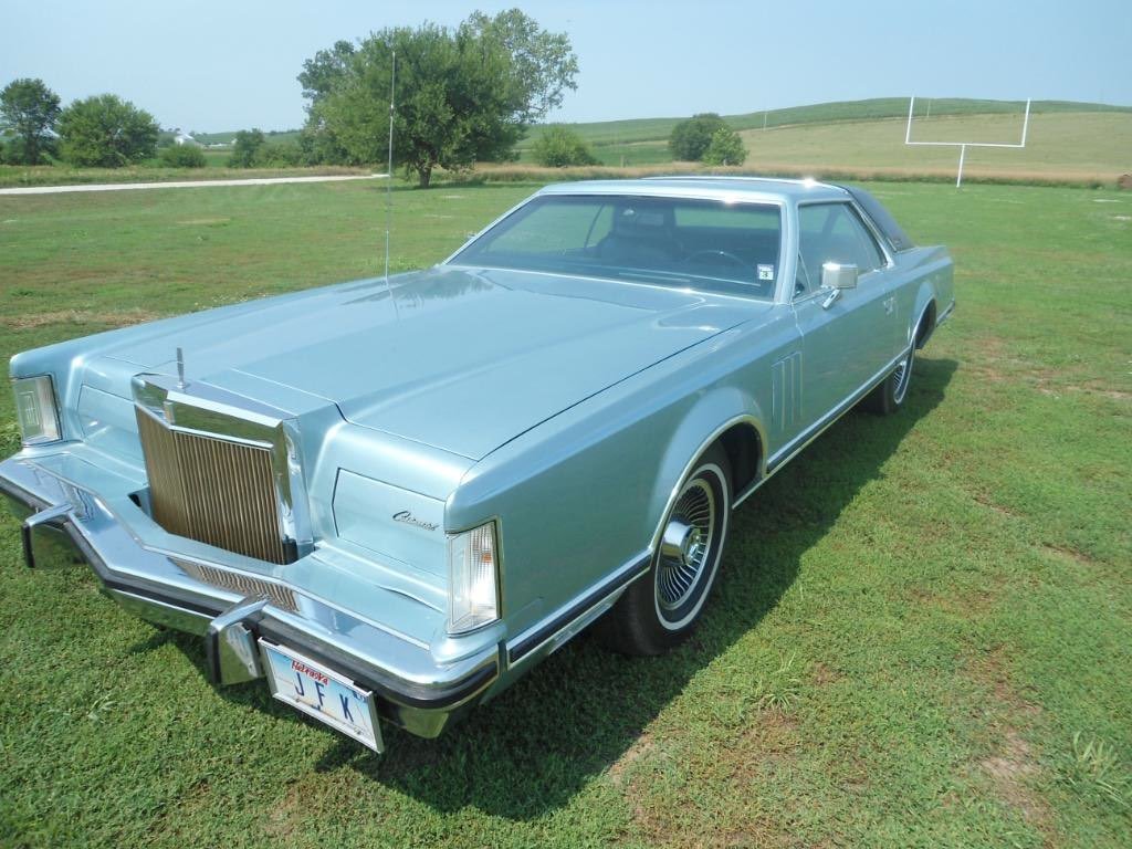 A classic light blue 1970s Chrysler car parked on grass in a rural field with goalposts in the background.