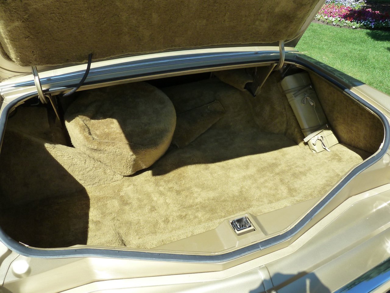 The open trunk of a vintage beige car lined with tan carpet, showing a spare tire and small beige bag inside.