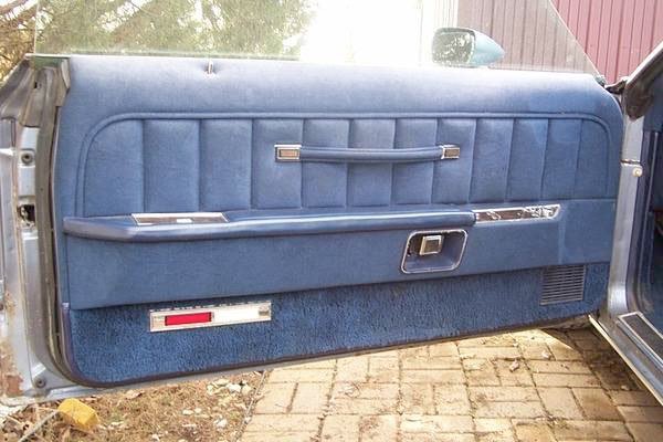 The interior side of a vintage blue car door with a fabric panel, handle, and storage compartments.