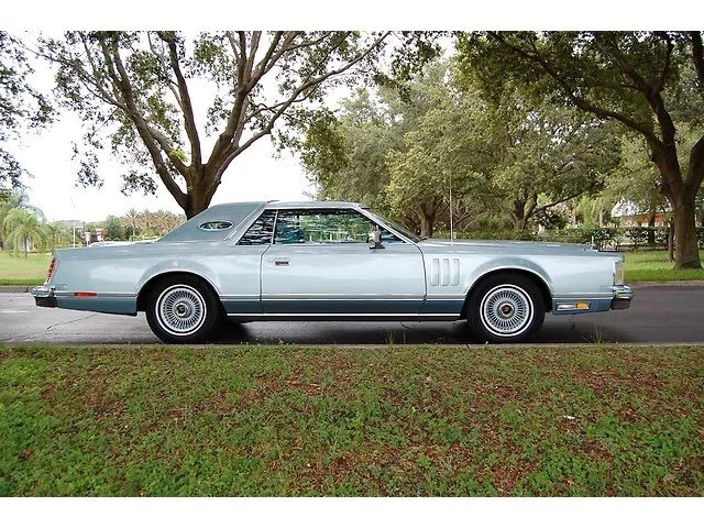 A vintage silver luxury car parked on a street with trees and greenery in the background.