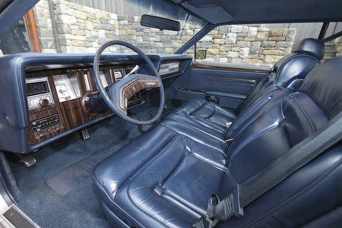 Interior of a vintage car showing blue leather seats, a large wooden steering wheel, and a dashboard with wood trim, parked against a stone wall.