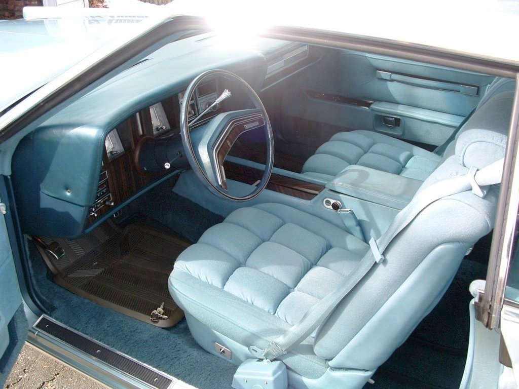The interior of a vintage car showing the front seats, dashboard, steering wheel, and part of the rear seat, all in a light blue color with wood trim accents.