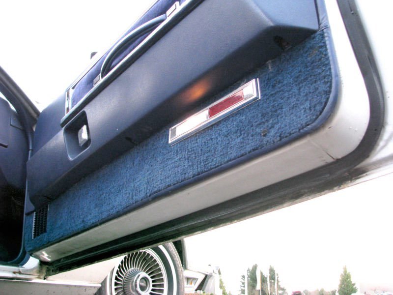 View of the interior of a classic car, showing the underside of the dashboard and the footwell area with blue carpeting and a rectangular light or reflector. Part of the front wheel and some trees are visible outside.