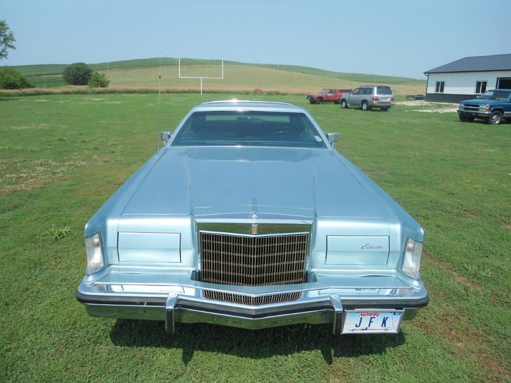 A vintage silver Mercury Cougar parked on a grassy field with a racetrack in the background and several other cars nearby.