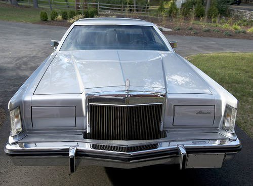 Front view of a vintage silver luxury car, likely a classic model from the 1970s, parked outdoors on a driveway with trees and grass in the background.