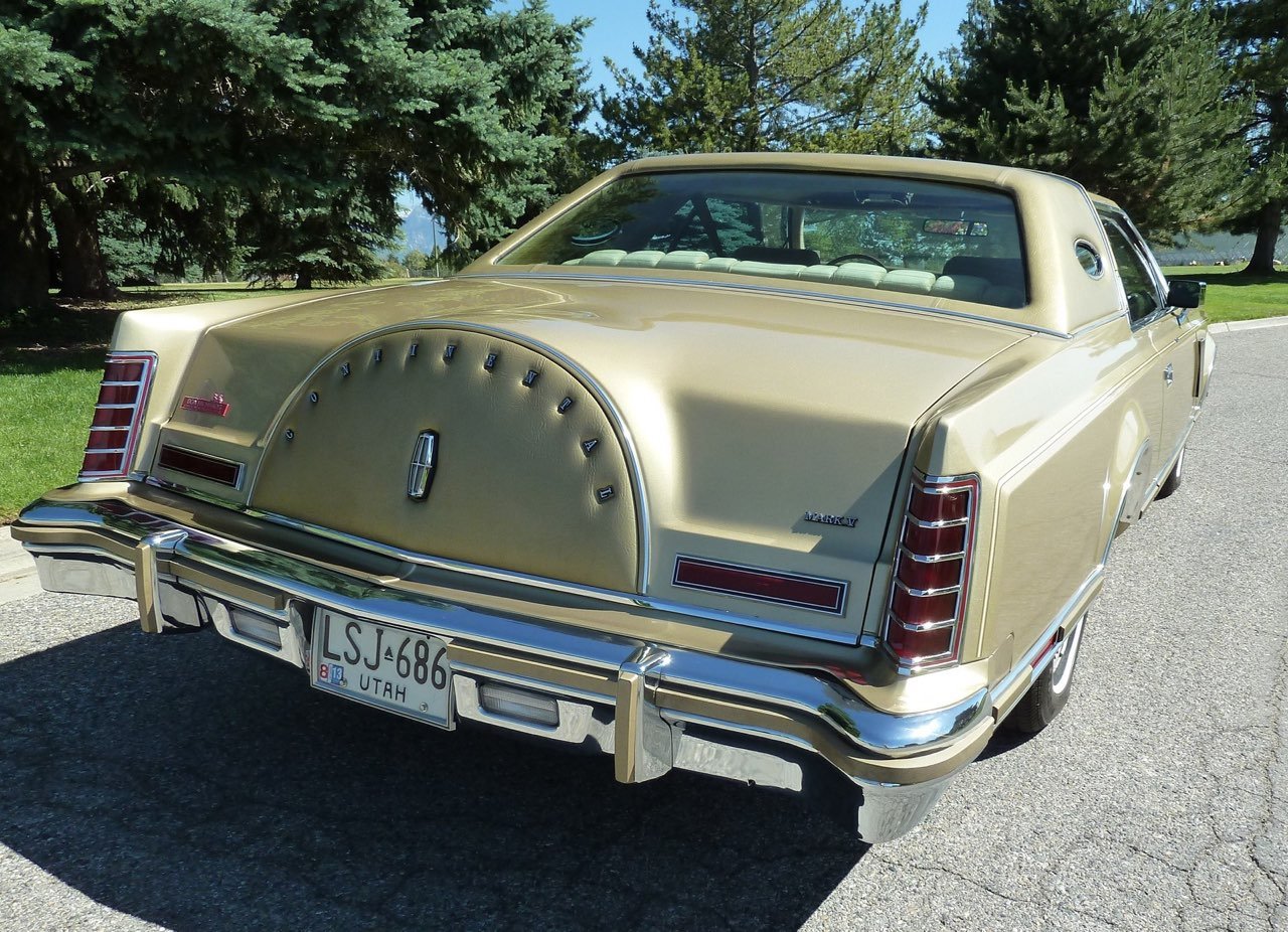 A vintage Lincoln Mark IV car in beige color parked on a paved road with green trees in the background.