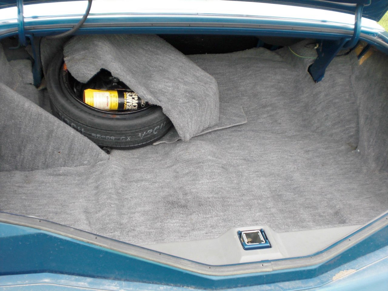 The trunk of a car with gray carpeting, containing a spare tire, a bottle of car fluid, and some gray carpeting pieces.