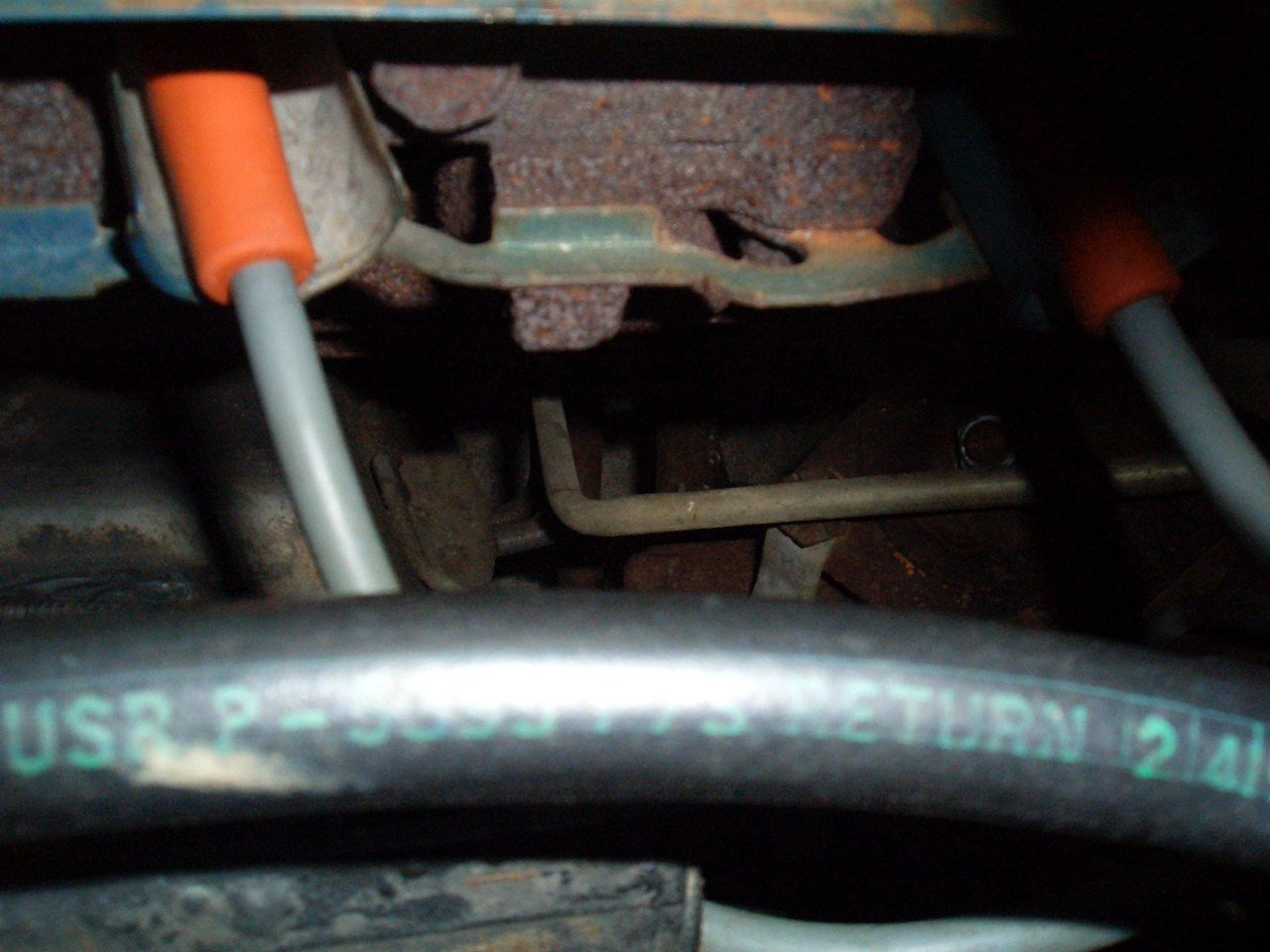 Close-up of engine components showing rusty metal parts, hoses, and spark plug wires in a car engine bay.