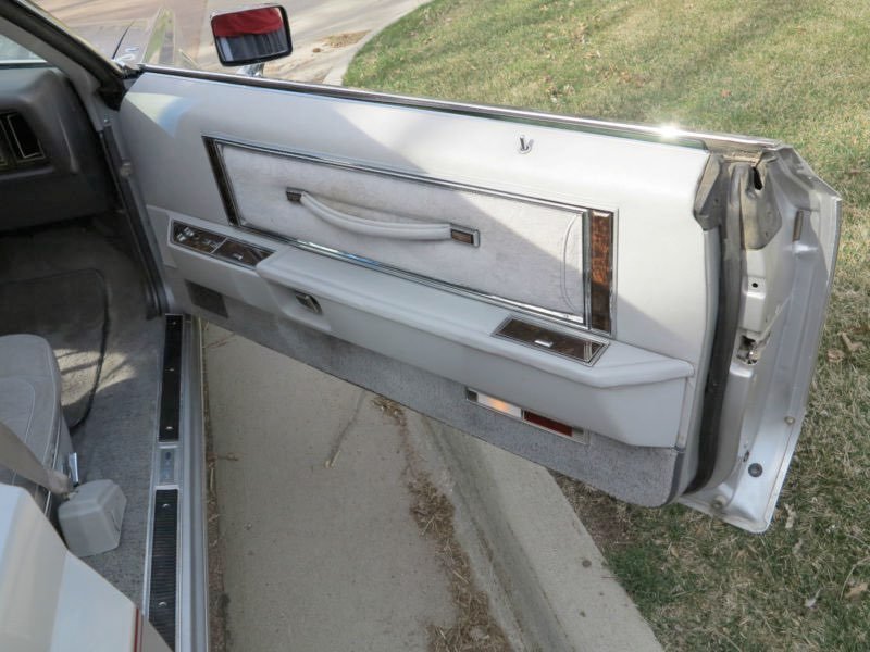 Close-up of a vintage car door interior with light gray and wood trim, fabric-covered panel, and manual window crank.