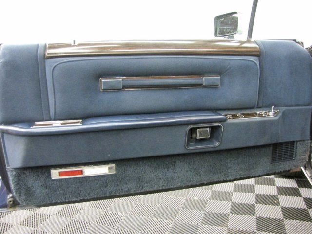 Close-up of a vintage car's interior dashboard, showing blue upholstery, a horizontal chrome handle, and a carpeted lower panel with a small vent.