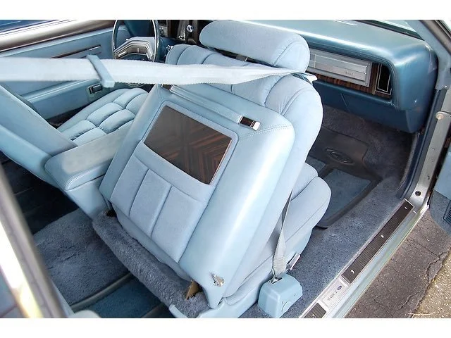 Interior of a vintage car with a light blue leather backseat, plush headrests, and wood trim. The seat has a built-in screen, and the seatbelt is visible across the seat.