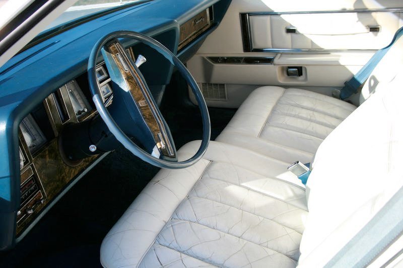 Inside the front cabin of a vintage car, featuring a white leather seat, a large steering wheel with a wood and metal trim, and a dashboard with various gauges and controls.
