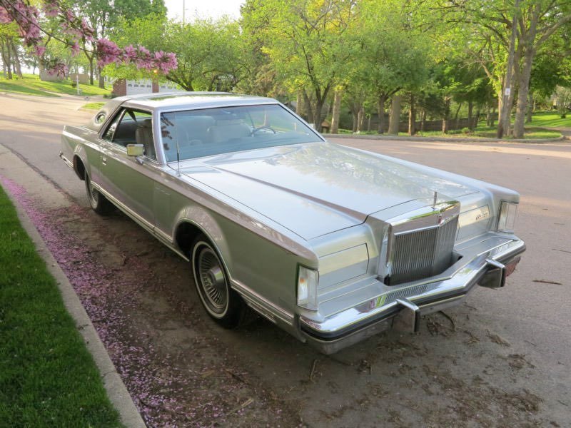 A vintage silver luxury car parked on a street with trees and pink flowers nearby.