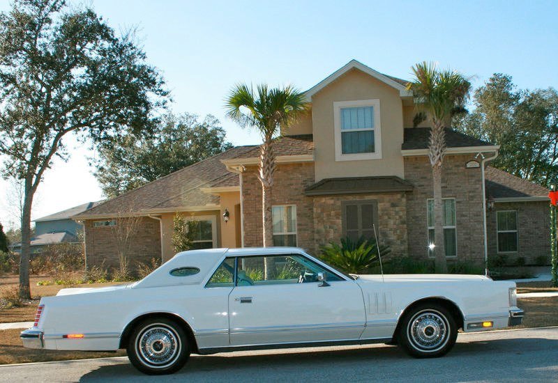 A classic white car parked in front of a two-story house with palm trees.