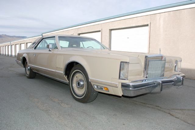 A beige vintage Cadillac sedan parked on a rooftop parking lot with garages in the background.