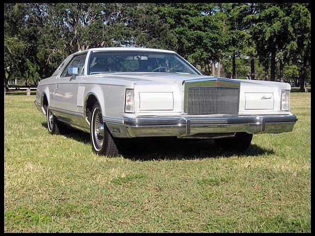 A vintage white luxury sedan parked on a grassy field with trees in the background.