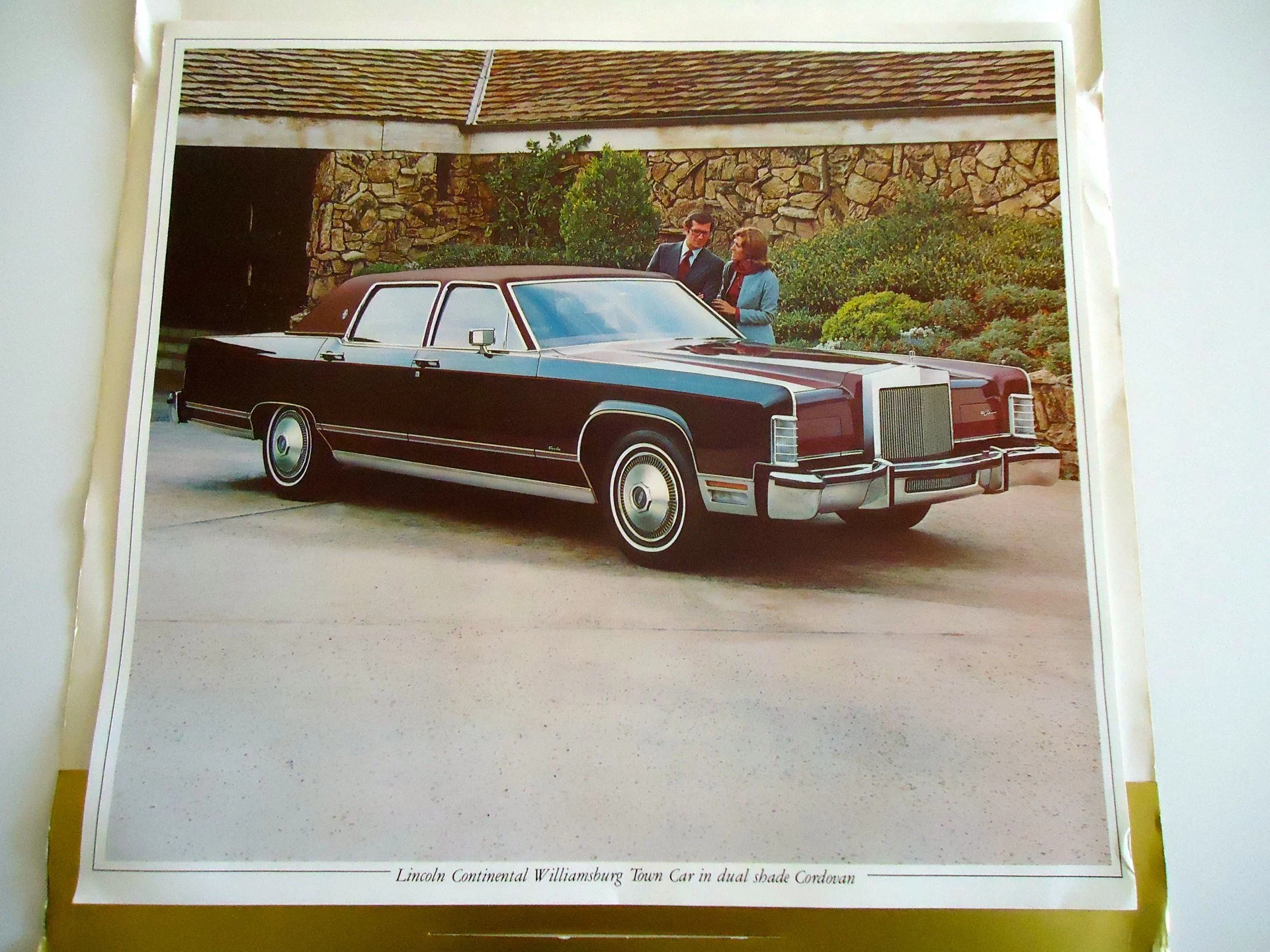 A vintage black Lincoln Continental Westminster town car with dual shade Cordovan, parked outside with two people in business attire standing near it. The background includes a stone wall and greenery.