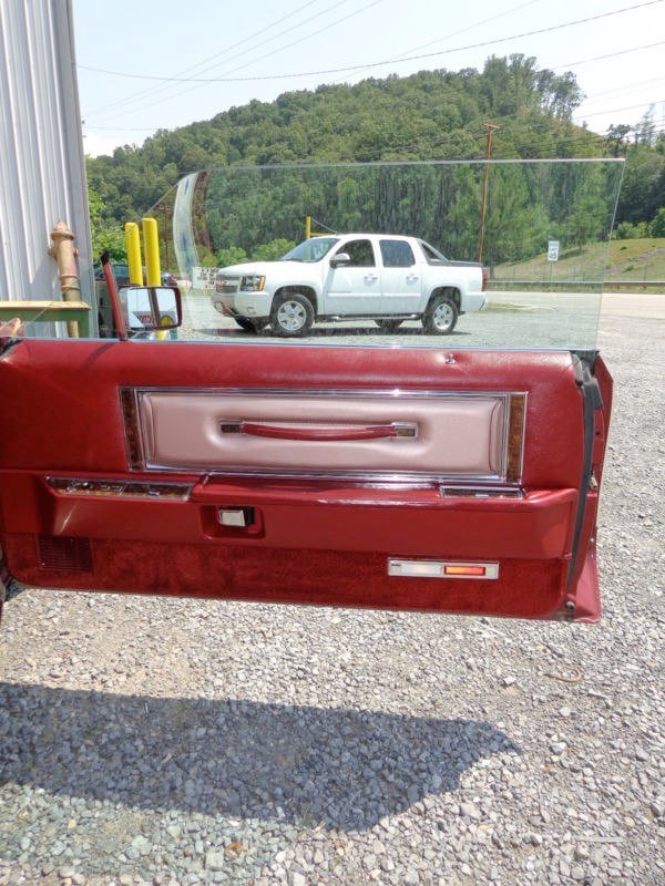 Inside view of a red vehicle door with a beige interior panel, handle, and control panel, with a clear glass window and a white pickup truck parked in the background on a gravel lot, with green hills, trees, and cloudy sky.