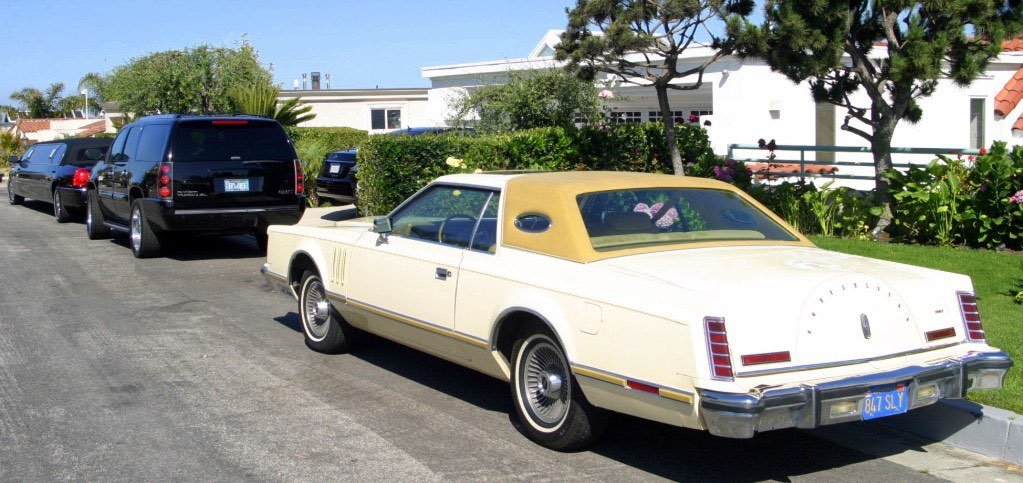 A white vintage convertible car with a beige top parked on a residential street, with other modern vehicles and a house with a garden in the background.