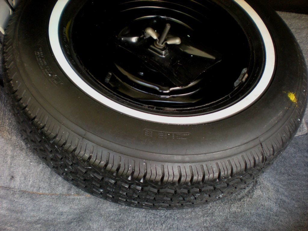 A close-up of a black car tire with a white sidewall and a black wheel rim, resting on a textured gray surface.