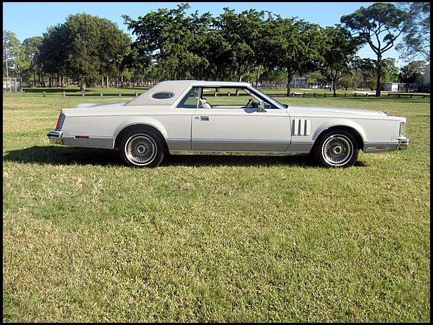 Silver vintage two-door car parked on grass with trees in the background.