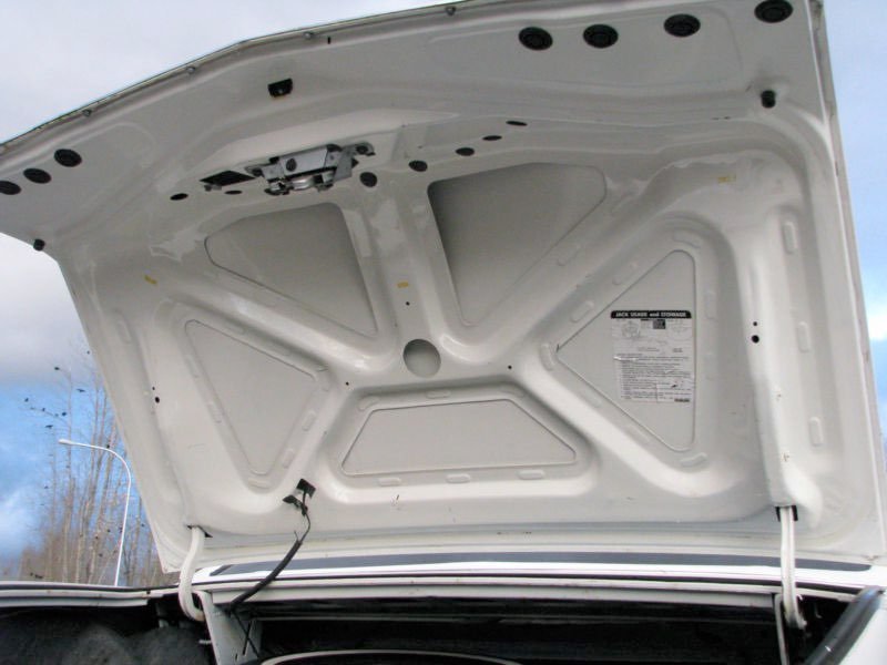 The underside of a white vehicle's hood, showing structural supports and a sticker with safety and maintenance information.