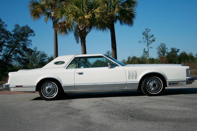 A vintage white luxury car parked on a paved road with palm trees and a clear blue sky in the background.