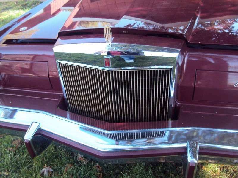 Close-up of the front of a vintage maroon car, showcasing a large chrome grille and bumper.