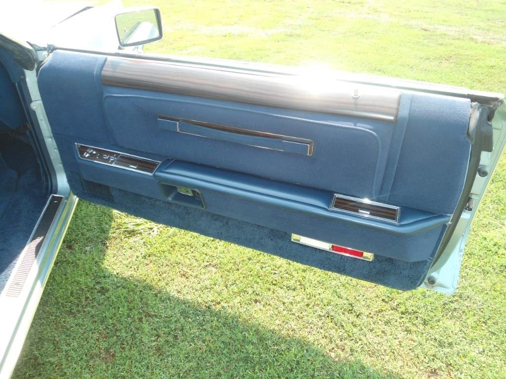 Close-up of a vintage car's interior door panel, made of blue fabric and plastic, with chrome accents and a wooden trim on top.