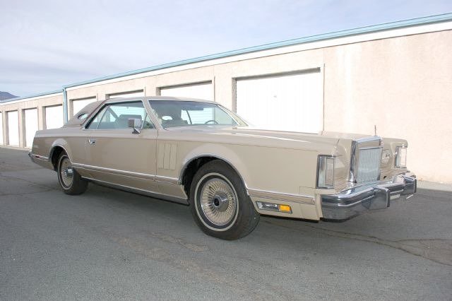 A vintage beige luxury car parked on the street in front of a beige building with garage doors.