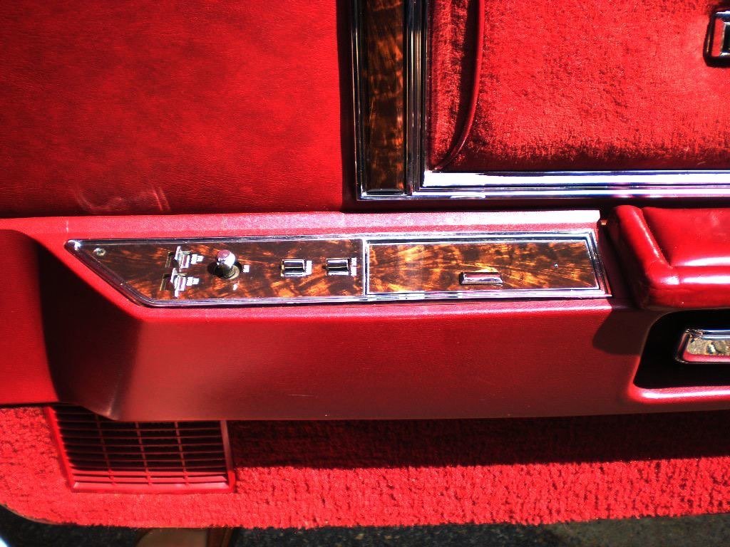 Close-up of the interior of a vintage car door panel with control switches and wood trim, featuring red upholstery and carpet.