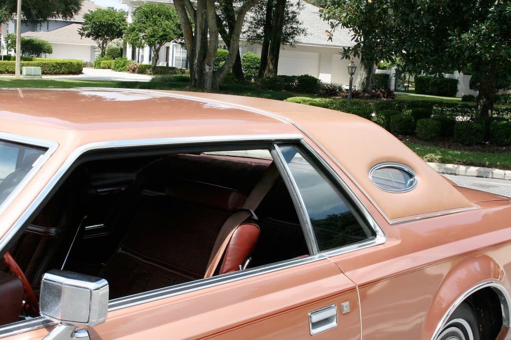 Close-up of a vintage peach-colored car parked outdoors, showing the side window, side mirror, and part of the roof with a decorative oval vent.
