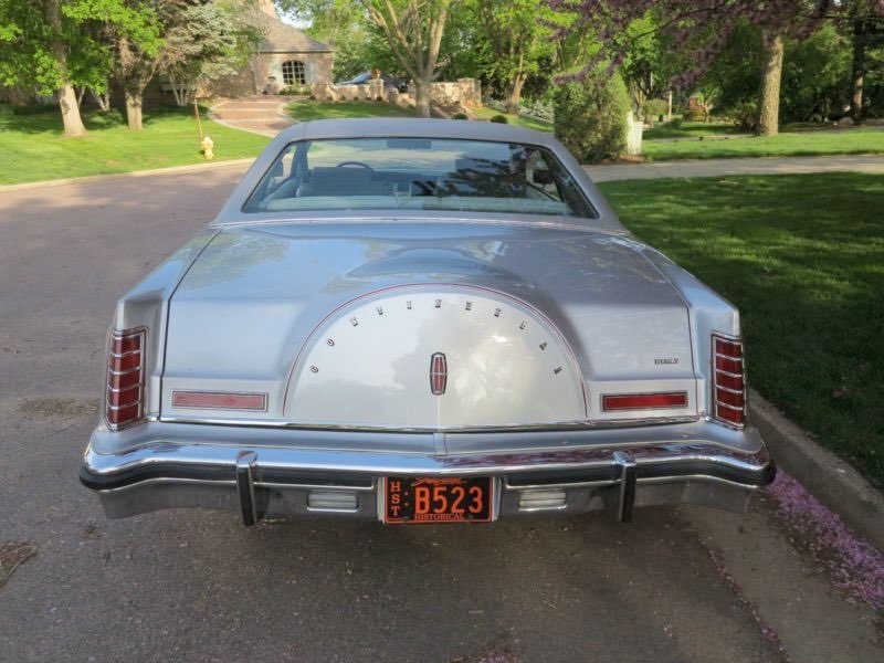 Rear view of a vintage Lincoln car with a distinctive rounded trunk design, parked on a residential street with trees and a house in the background.