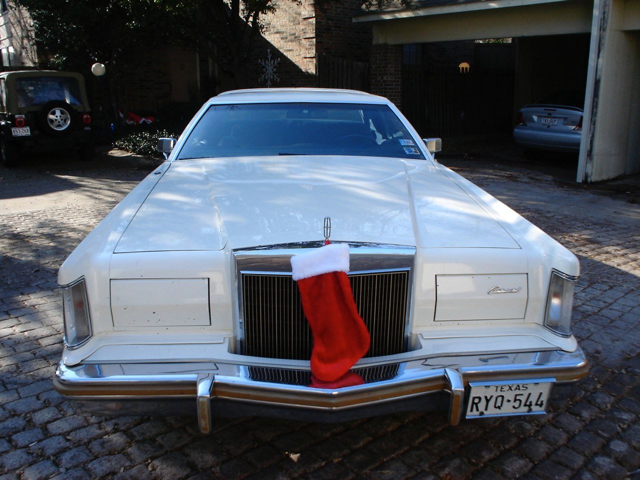 A white vintage Lincoln car decorated with a red Christmas stocking hanging on the front grille, parked on a brick driveway with Christmas ornaments and decorations nearby.