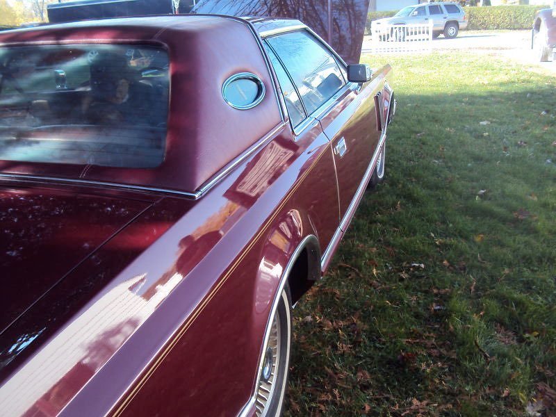 A vintage maroon car parked on grass, viewed from the rear passenger side, with reflections on its shiny surface and other parked vehicles in the background.