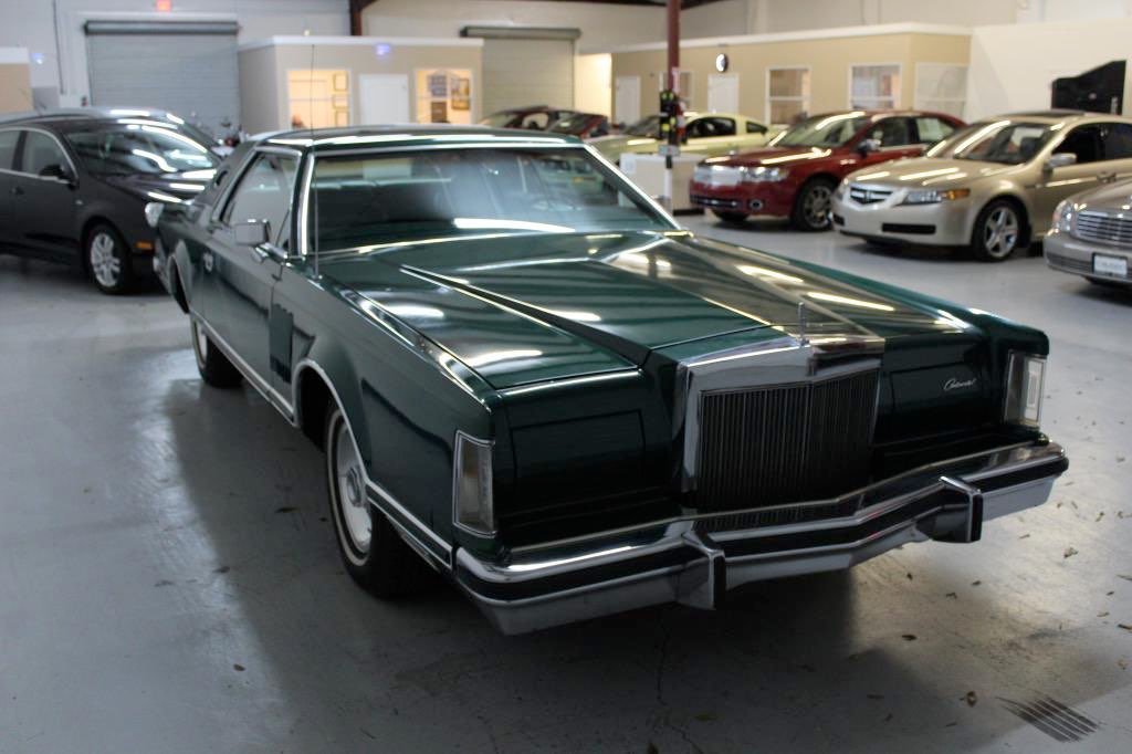 A vintage dark green Cadillac car inside an indoor showroom with other modern cars parked in the background.