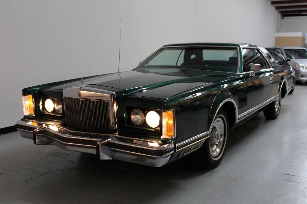 A vintage dark green classic car with chrome accents parked indoors, with other cars visible in the background.